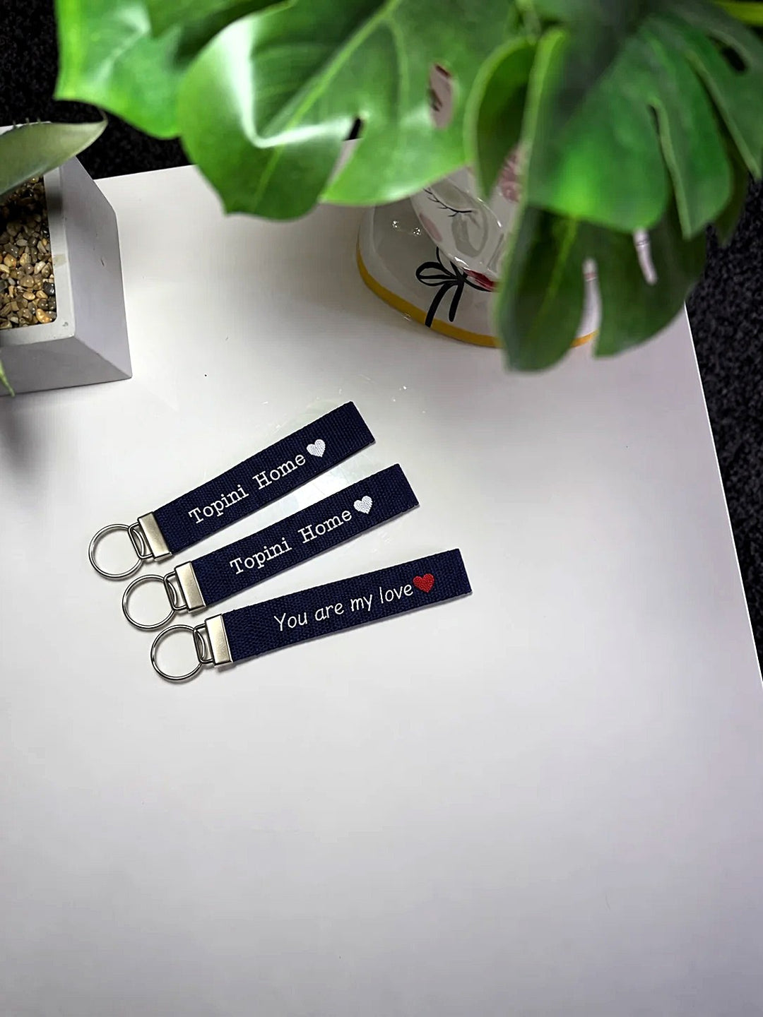 Tre portachiavi in tela blu navy con frase ricamata e cuore, su piano bianco